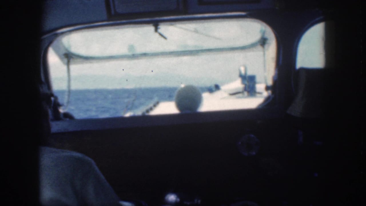 1959 Oahu 8mm frame showing a man in a white shirt seated inside a boat cabin, looking toward open water through the front window.