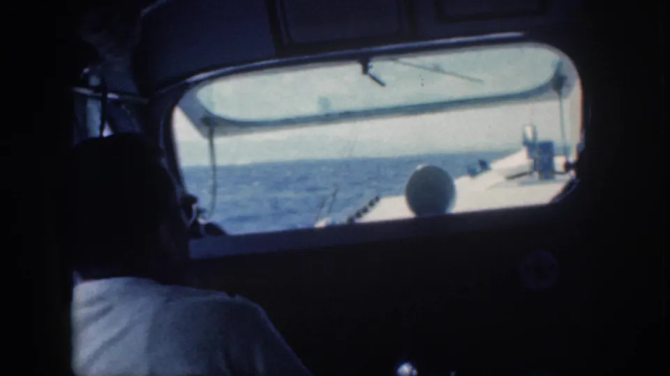 1959 Oahu 8mm footage frame with a seated man in a white shirt inside a small vessel, framed against ocean and daylight through the window.