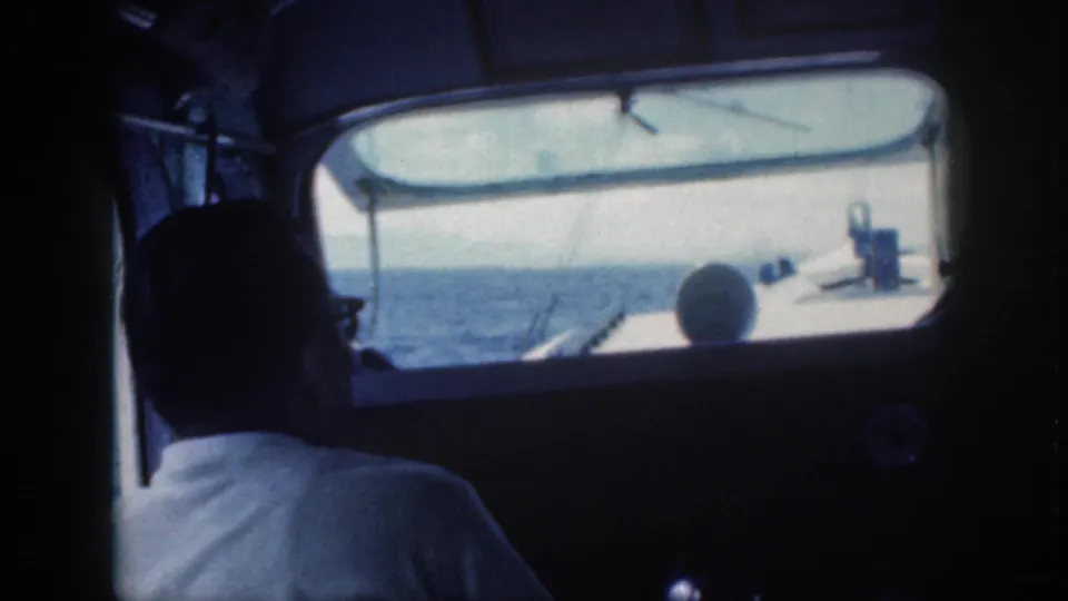 1959 Oahu frame showing a shadowed seated figure inside the boat cabin, with blue water, sky, and bow details visible ahead.