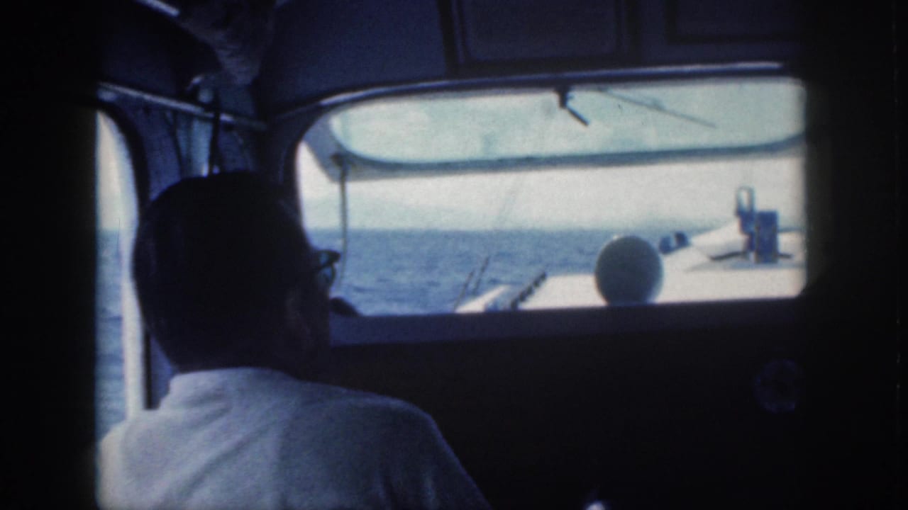 Inside a 1959 Oahu power boat cabin, a man in glasses sits in shadow while the open sea and bow area appear through the front window.