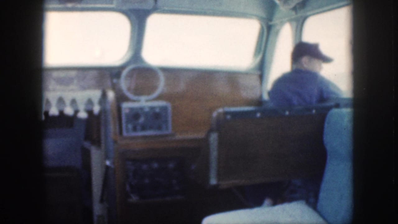 A wood-paneled cabin helm area on a 1959 Oahu power boat shows controls, a small circular mirror or fixture, and a man in a dark cap near the front windows.