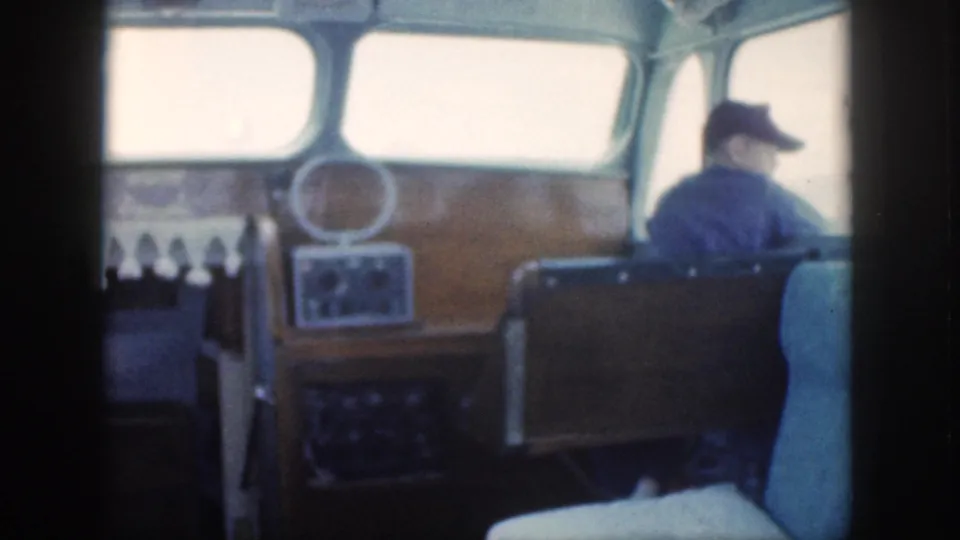 A wood-paneled cabin helm area on a 1959 Oahu power boat shows controls, a small circular mirror or fixture, and a man in a dark cap near the front windows.