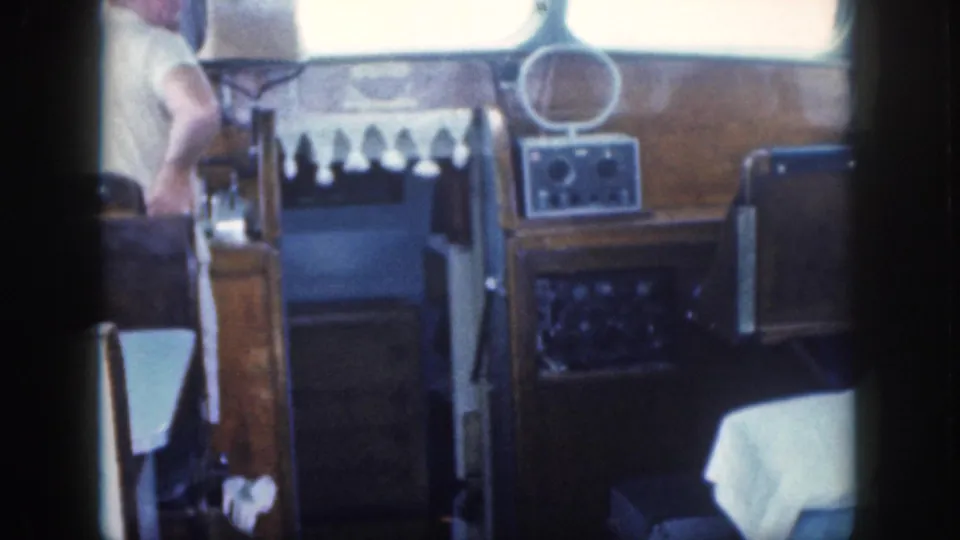 Inside the 1959 Oahu boat cabin, an adult man stands or moves beside the helm while the wood-paneled dashboard, controls, chair backs, and bright windows remain visible.