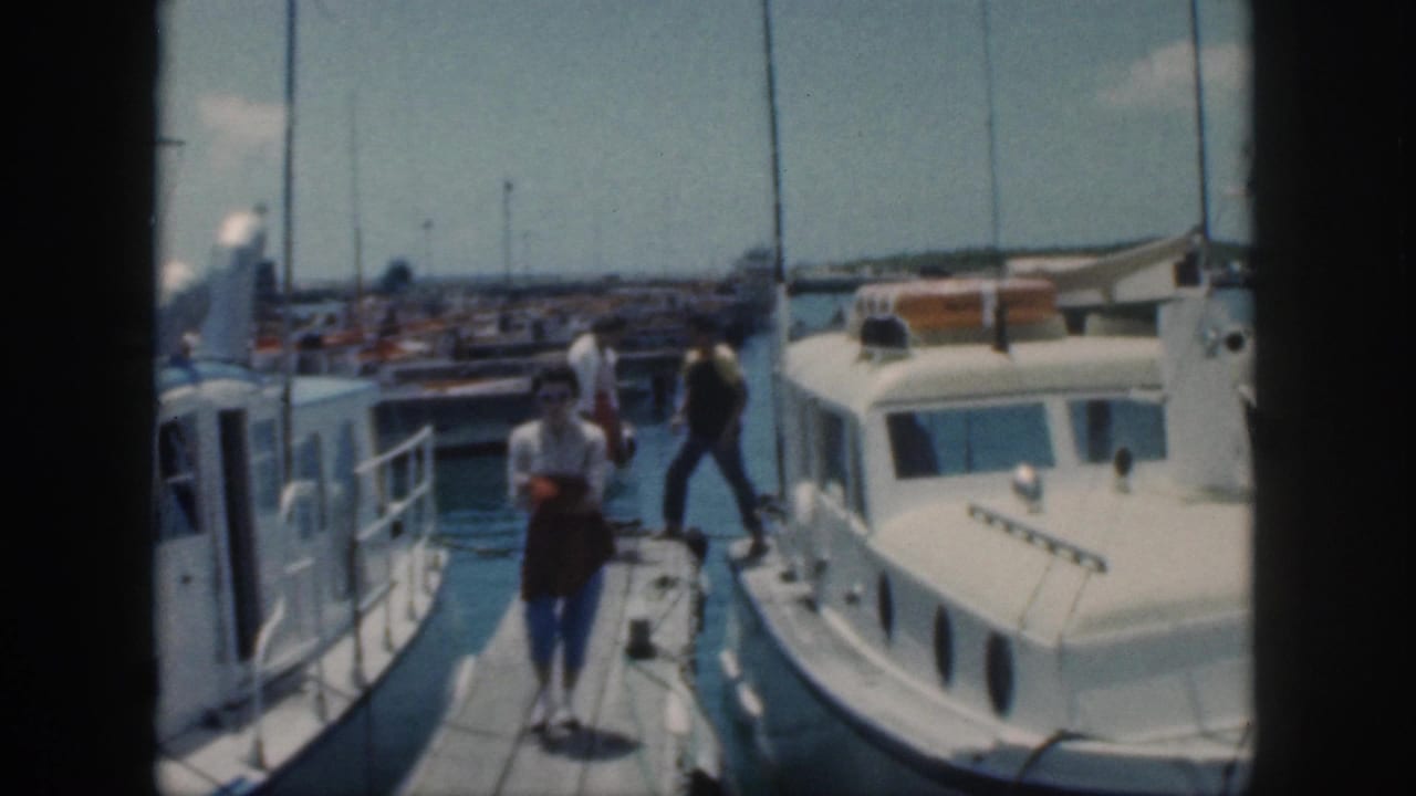 A woman walks toward the camera on a dock while others stand near boats.