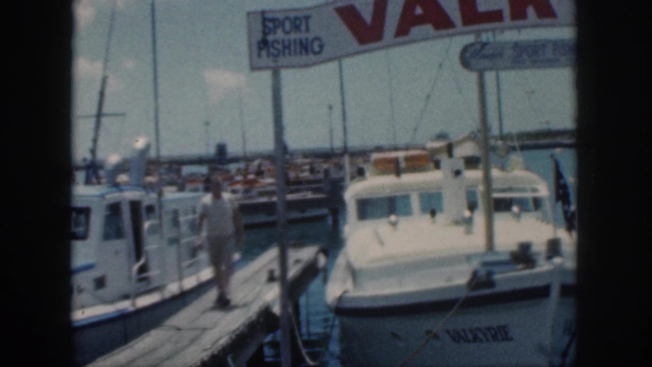 1959 Oahu home-movie frame showing a man on a wooden dock between boats, with Valkyrie moored under sport fishing signage.