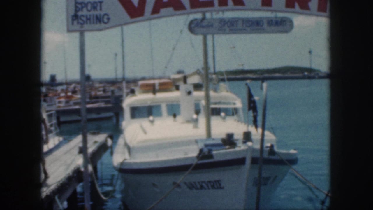 1959 Oahu frame of the white motorboat Valkyrie tied at a marina dock with calm water and distant shoreline behind it.