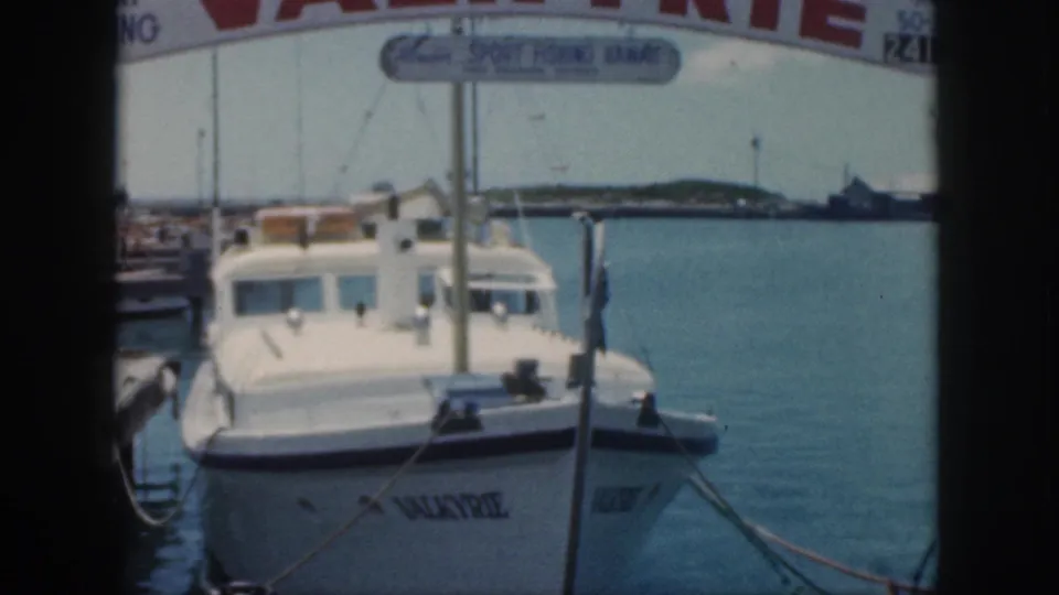 1959 Oahu marina frame showing Valkyrie tied to the dock beneath partial sport fishing signage with open water beyond.