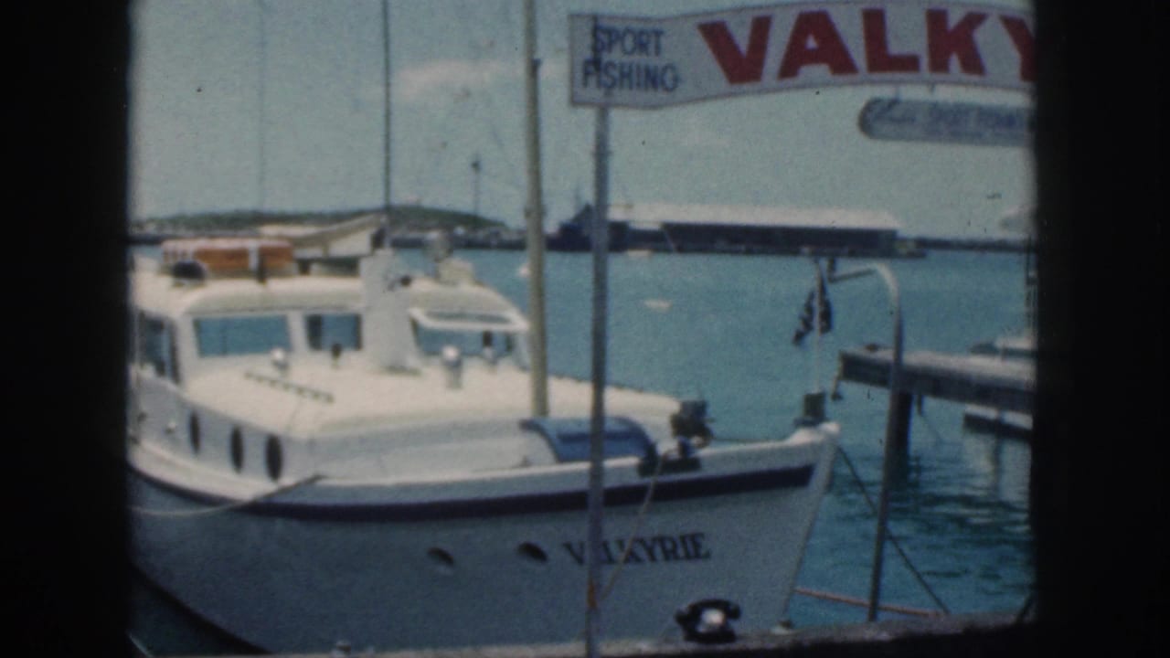 1959 Oahu home-movie frame of the white motorboat Valkyrie moored at a harbor beneath a sport fishing sign.