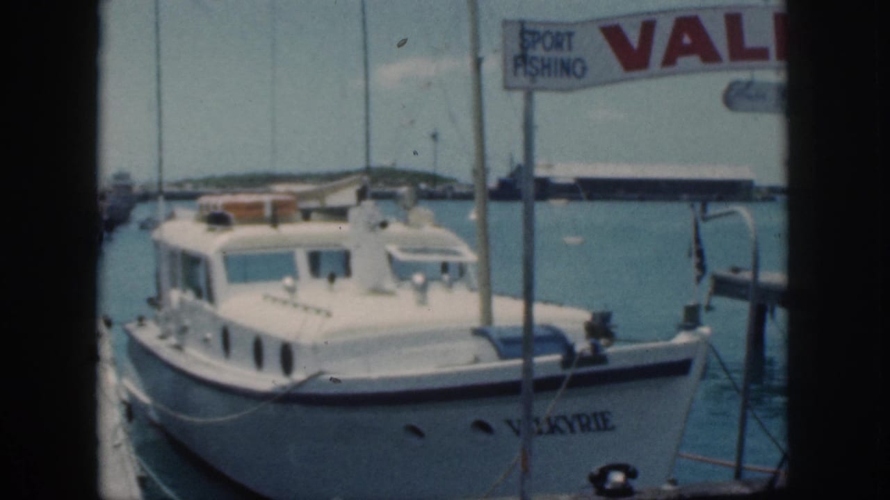 1959 Oahu harbor frame with the Valkyrie motorboat, dock posts, overhead sport fishing sign, and distant shoreline.
