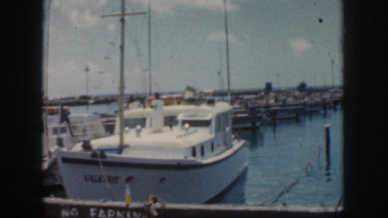 White cabin cruiser moored in a harbor with docks, pilings, and sailboat masts behind it.