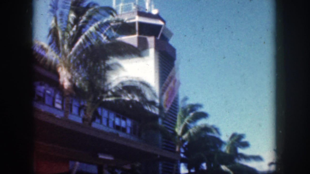 1959 Oahu airport terminal view with a control tower, palm fronds, blue sky, and dark 8mm film gate edges.