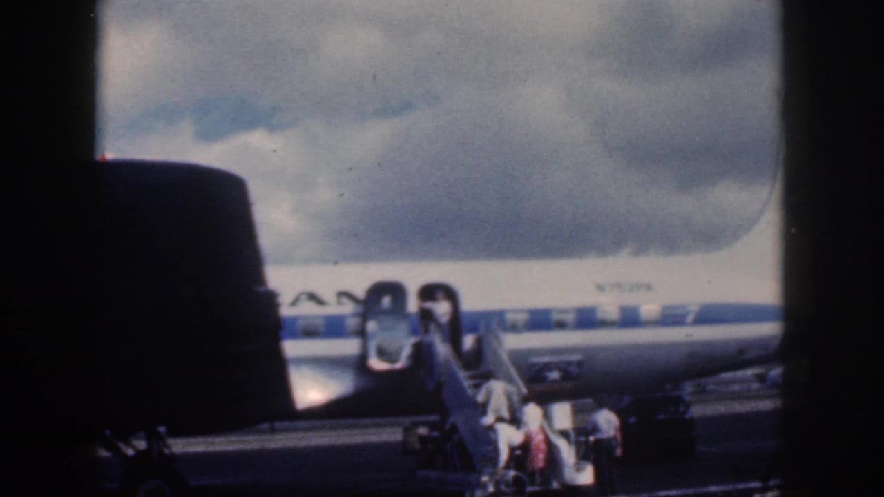 1959 Oahu home-movie frame showing passengers boarding an American Airlines propeller aircraft from mobile stairs under cloudy skies.