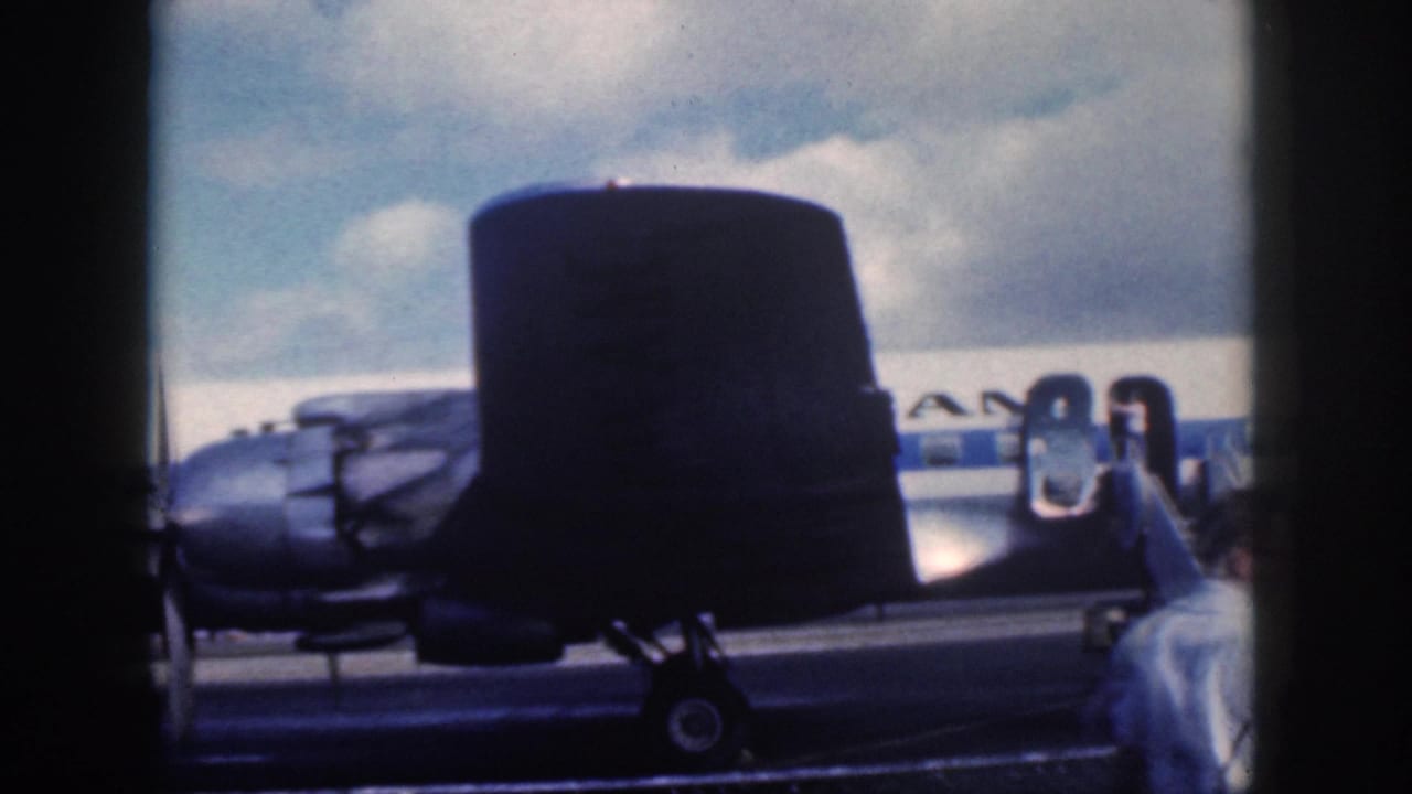 1959 Oahu airfield frame with a dark tail in the foreground and a passenger ascending mobile stairs to a propeller aircraft.