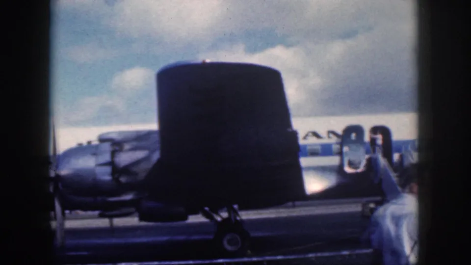 1959 Oahu airfield frame with a dark tail in the foreground and a passenger ascending mobile stairs to a propeller aircraft.