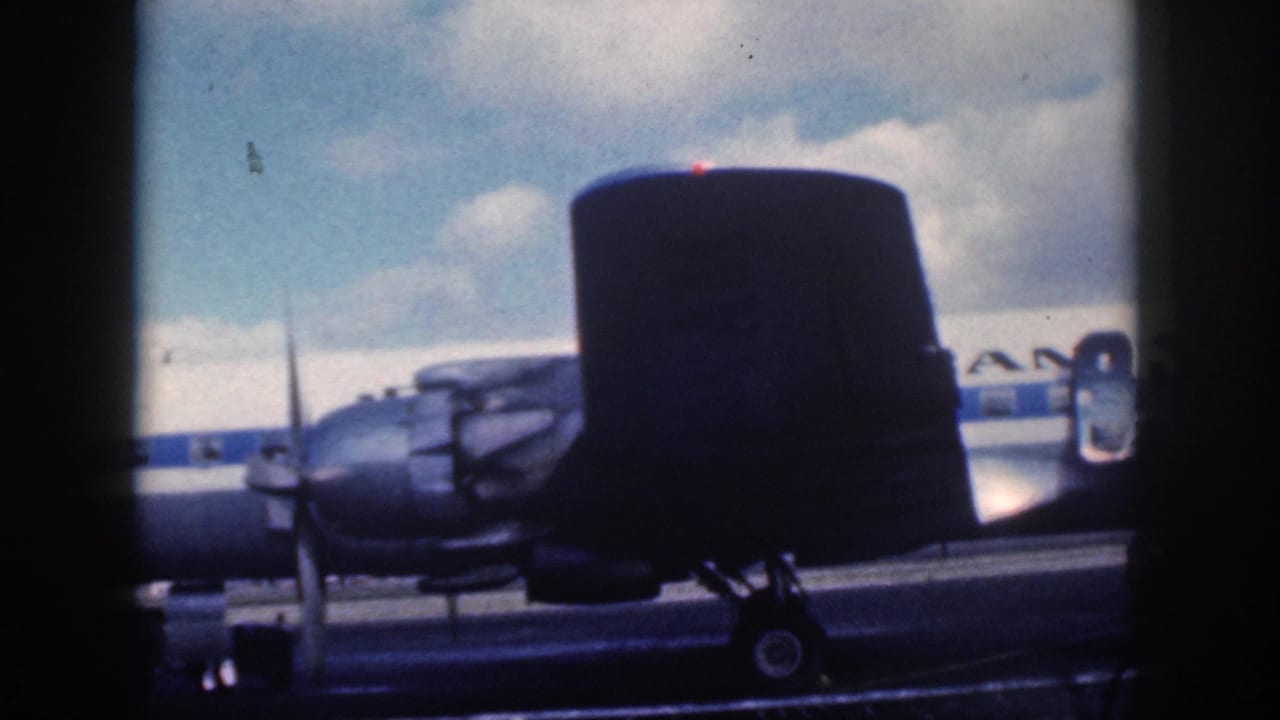 1959 Oahu home-movie frame showing a nearby aircraft tail in front of an American Airlines propeller plane on the tarmac.