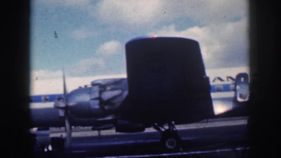 1959 Oahu home-movie frame showing a nearby aircraft tail in front of an American Airlines propeller plane on the tarmac.
