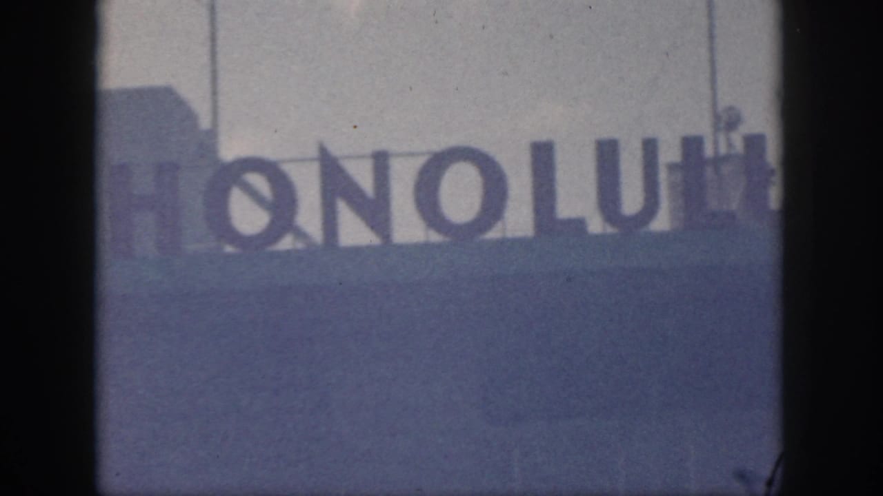 1959 Oahu home-movie frame showing partial dark letters from a Honolulu airport sign against a pale sky.
