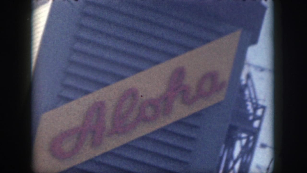 1959 Oahu home-movie view of the Aloha sign at an angle, with a blue wall and metal support structure beside it.
