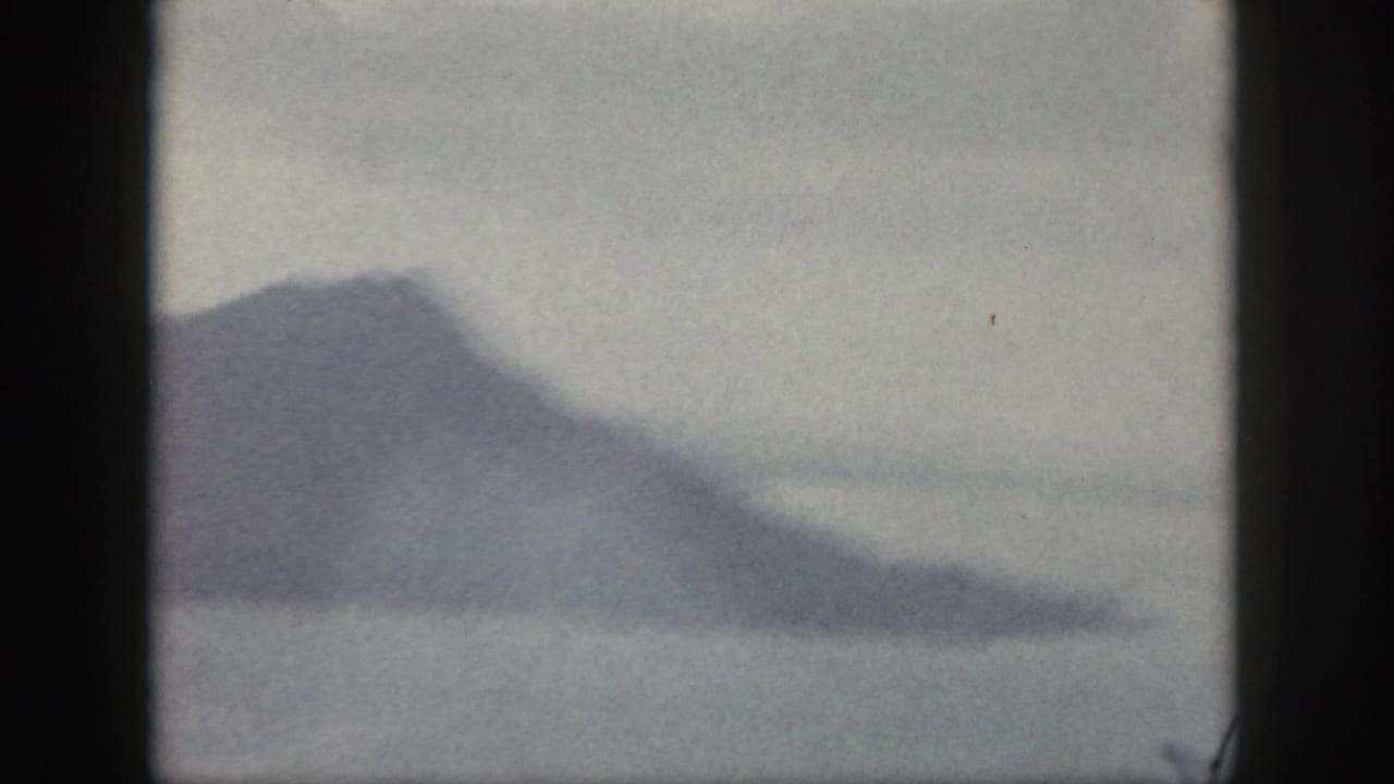 Hazy 1959 Oahu home-movie frame showing a dark mountain coastline under a pale, overcast sky.