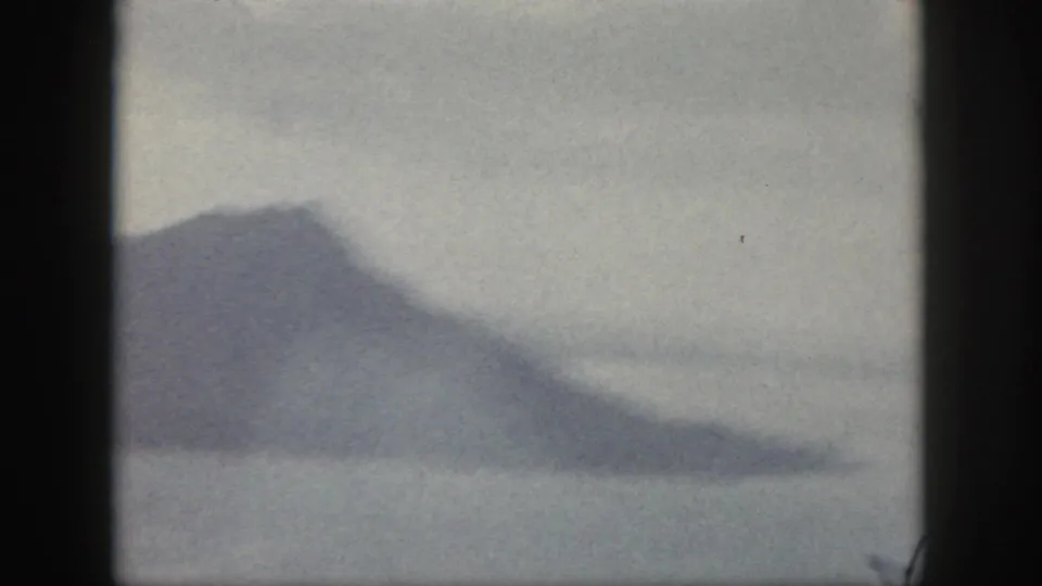 Hazy 1959 Oahu home-movie frame showing a dark mountain coastline under a pale, overcast sky.