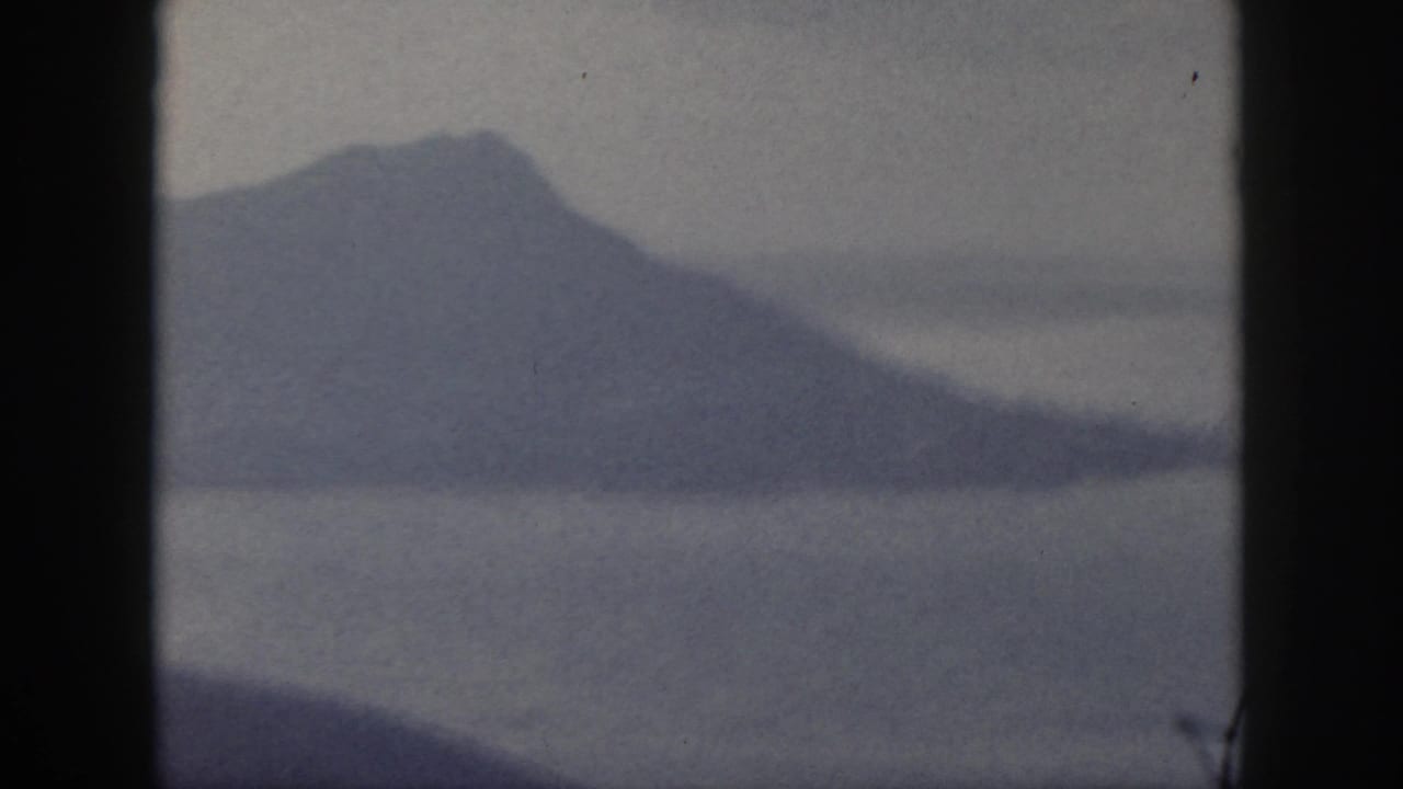 1959 Oahu home-movie frame showing a broad hazy mountain slope across calm water.