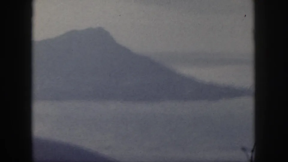 1959 Oahu home-movie frame showing a broad hazy mountain slope across calm water.