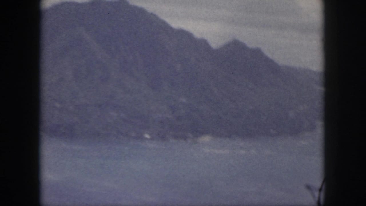 1959 Oahu 8mm frame with a broad blue-gray mountain ridge, pale sky, and water below.