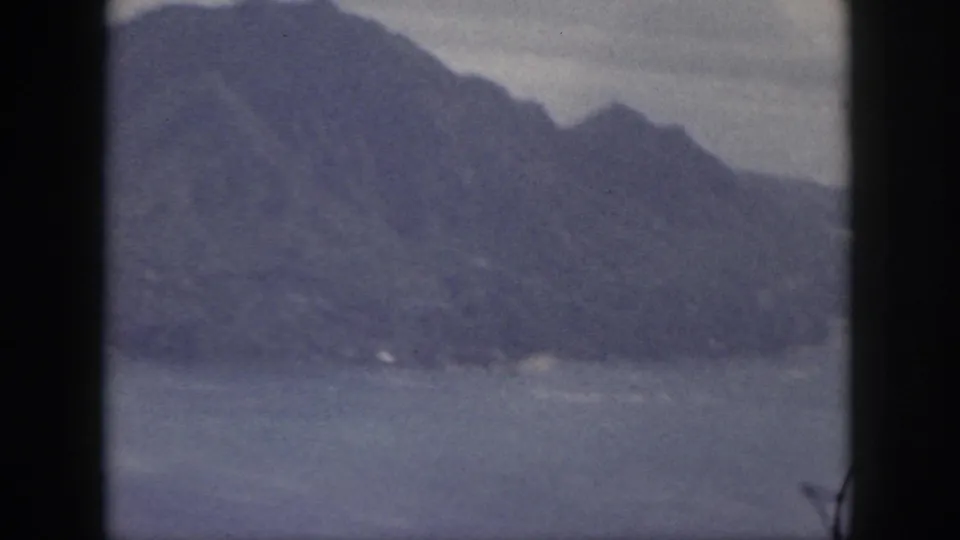 1959 Oahu 8mm frame with a broad blue-gray mountain ridge, pale sky, and water below.