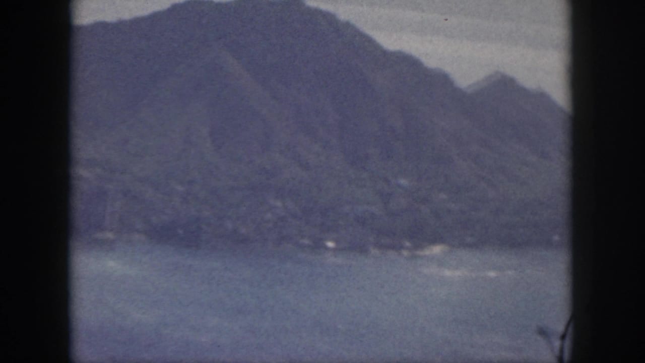 1959 Oahu home-movie frame showing coastal water and rugged mountains under a hazy sky, with visible 8mm frame borders.