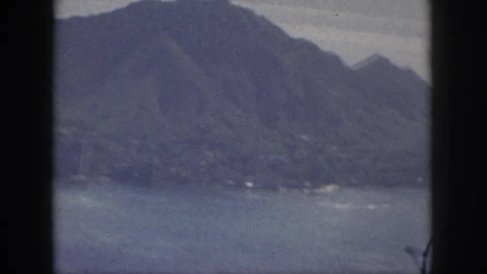 1959 Oahu home-movie frame showing coastal water and rugged mountains under a hazy sky, with visible 8mm frame borders.