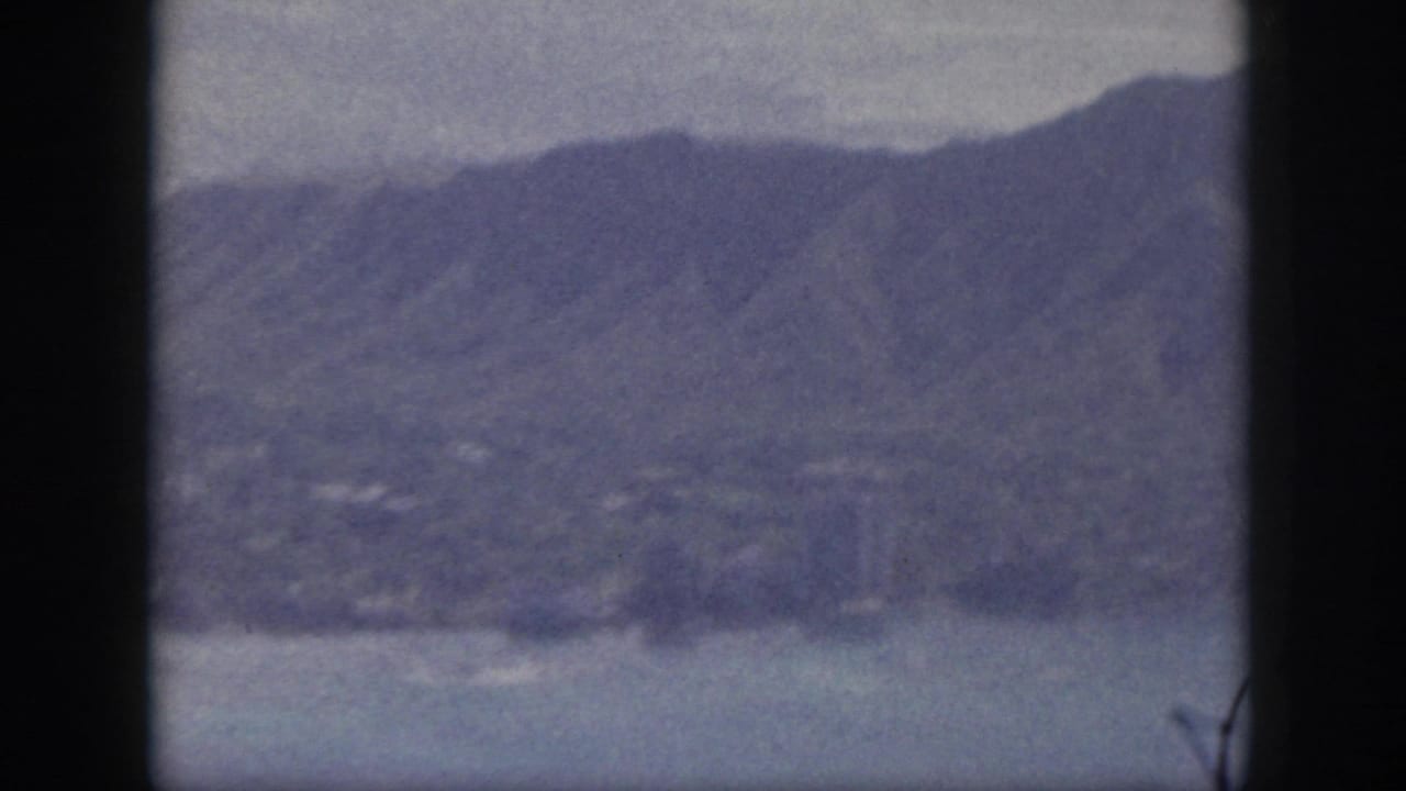 1959 Oahu aerial home-movie frame showing mountains, coastal water, shoreline, and faint distant settlement patterns.