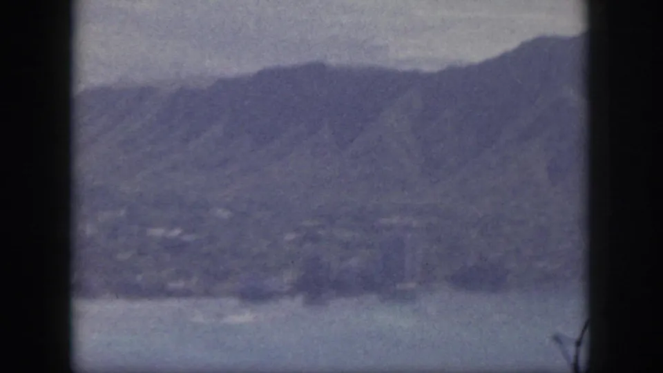 1959 Oahu aerial home-movie frame showing mountains, coastal water, shoreline, and faint distant settlement patterns.