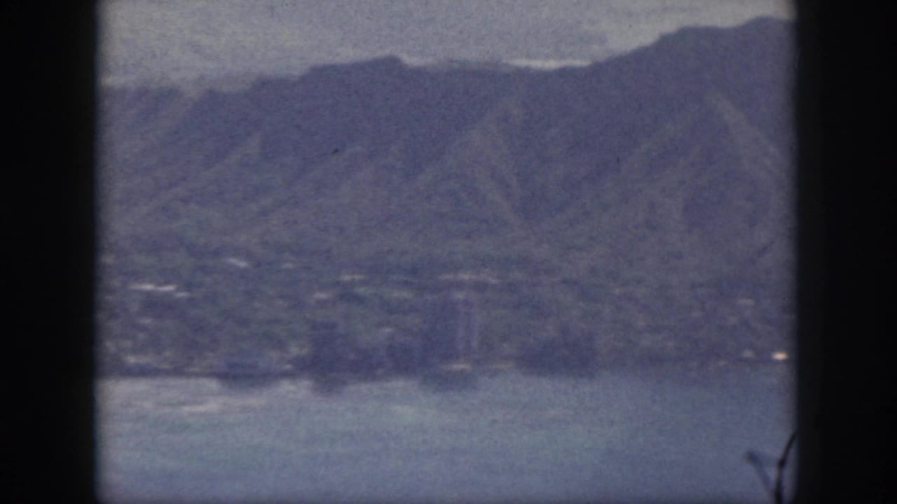 1959 Oahu aerial frame with mountain slopes, coastal water, shoreline, and faint structures visible through blue-tinted 8mm grain.