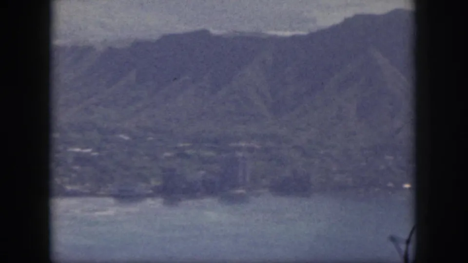 1959 Oahu aerial frame with mountain slopes, coastal water, shoreline, and faint structures visible through blue-tinted 8mm grain.