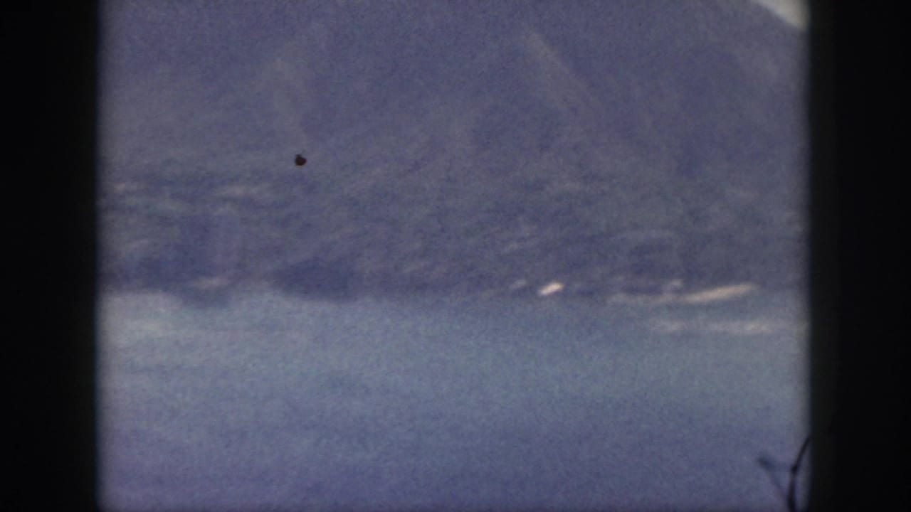 1959 Oahu home-movie view of hazy mountains beyond calm ocean water, framed by dark film gate edges.