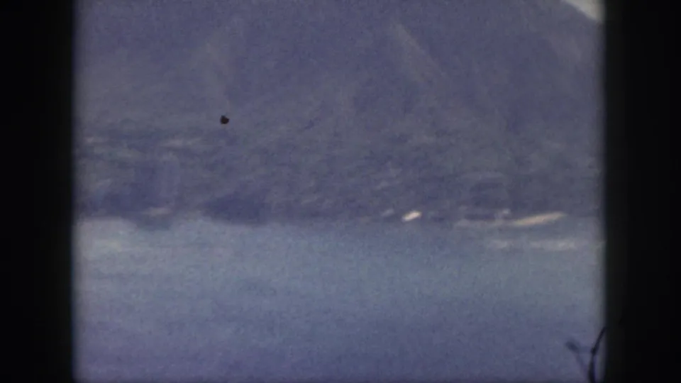 1959 Oahu home-movie view of hazy mountains beyond calm ocean water, framed by dark film gate edges.