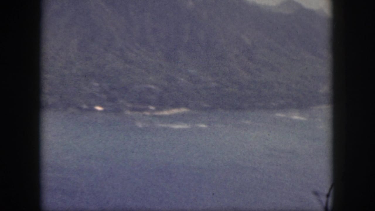 Soft 8mm frame of Oahu coastline with blue water, distant mountains, and a small bright shoreline detail.