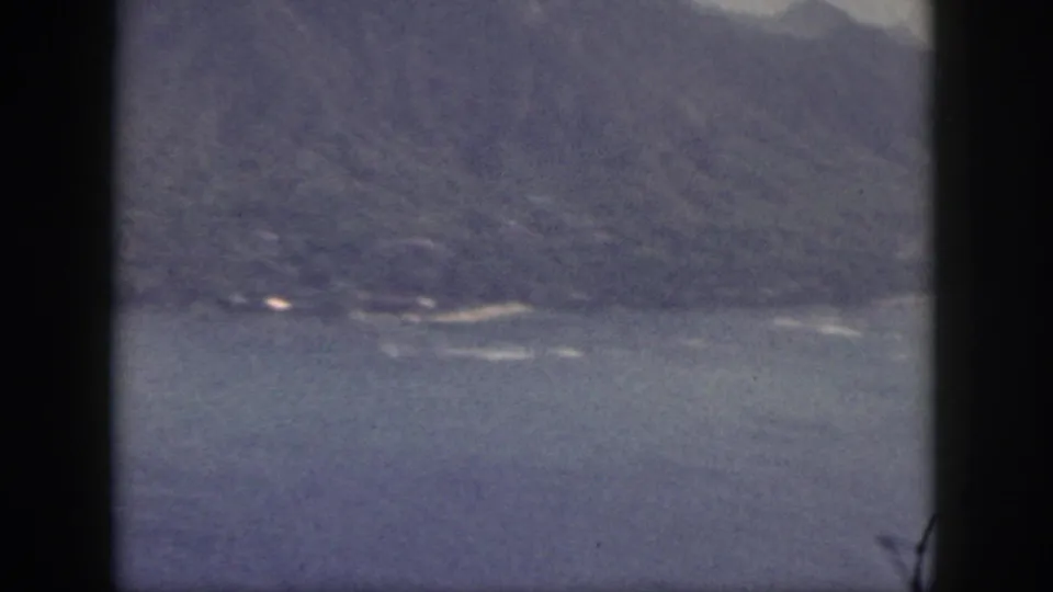Soft 8mm frame of Oahu coastline with blue water, distant mountains, and a small bright shoreline detail.