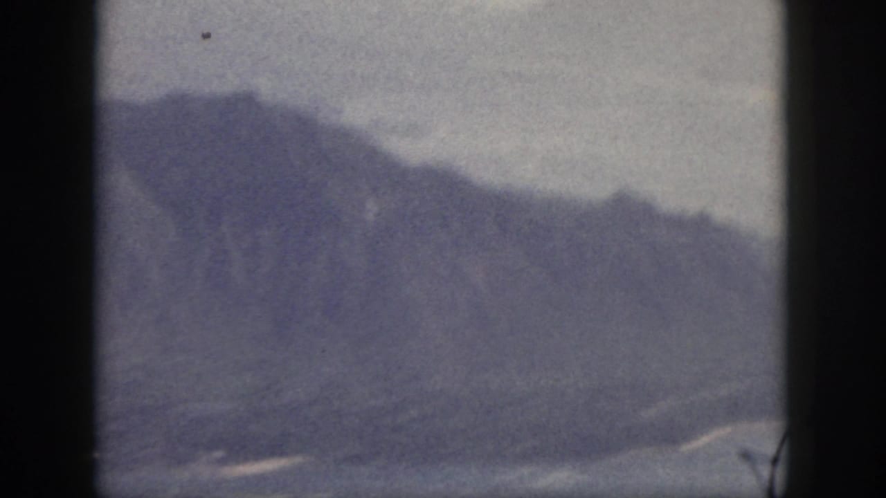 Mid-century Oahu 8mm view showing layered mountain slopes descending toward water under a pale sky.