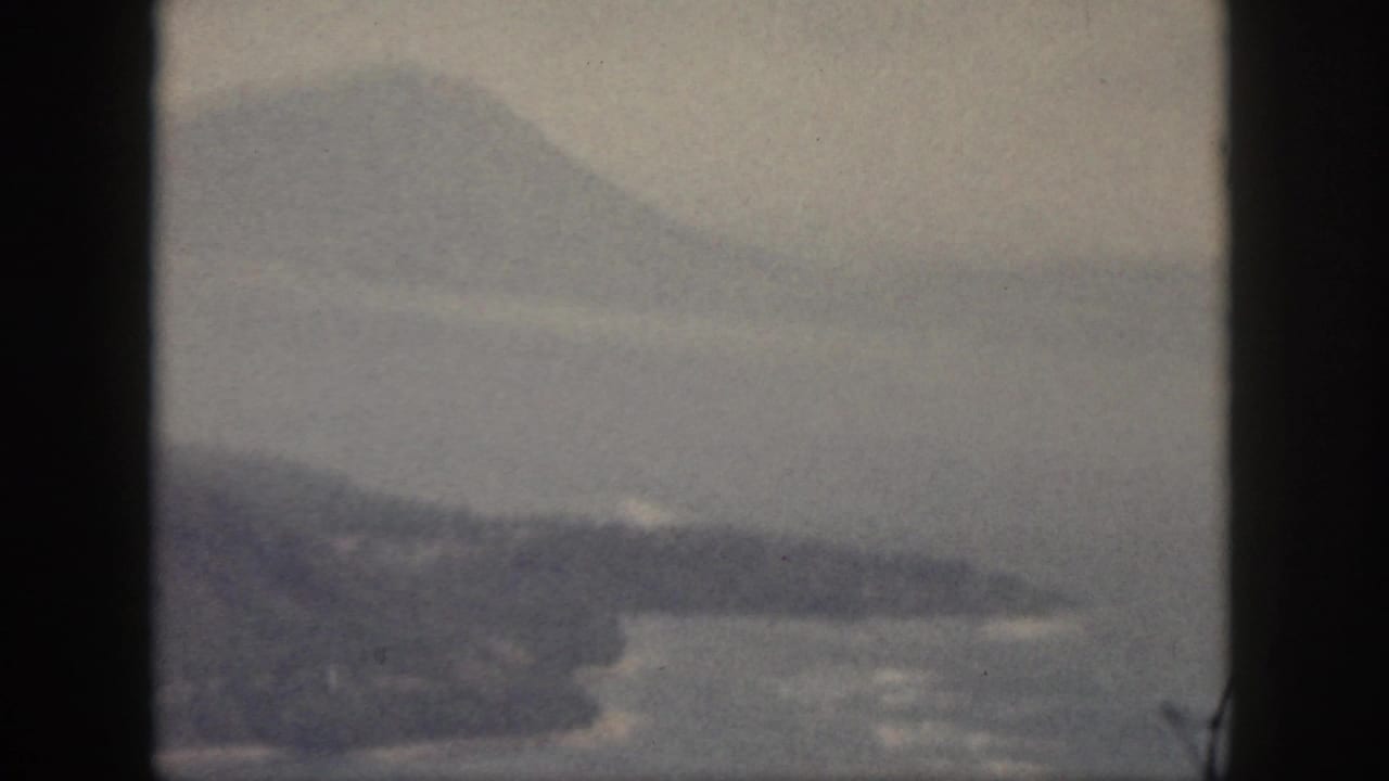 1959 Oahu home-movie frame of a low landform or island bank in calm water, with hazy mountains beyond.