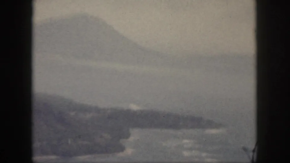 1959 Oahu home-movie frame of a low landform or island bank in calm water, with hazy mountains beyond.