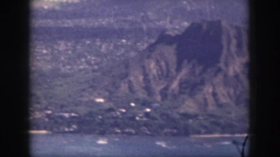 1959 Oahu 8mm view of a steep mountain slope, shoreline water, and scattered coastal buildings.