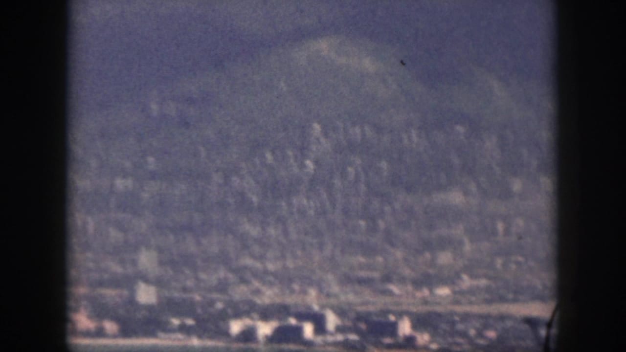 1959 Oahu home-movie frame showing a hazy coastal city and shoreline beneath forested mountains, viewed through dark frame bars.
