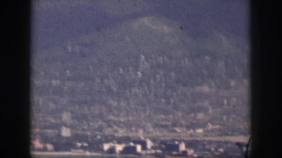 1959 Oahu home-movie frame showing a hazy coastal city and shoreline beneath forested mountains, viewed through dark frame bars.