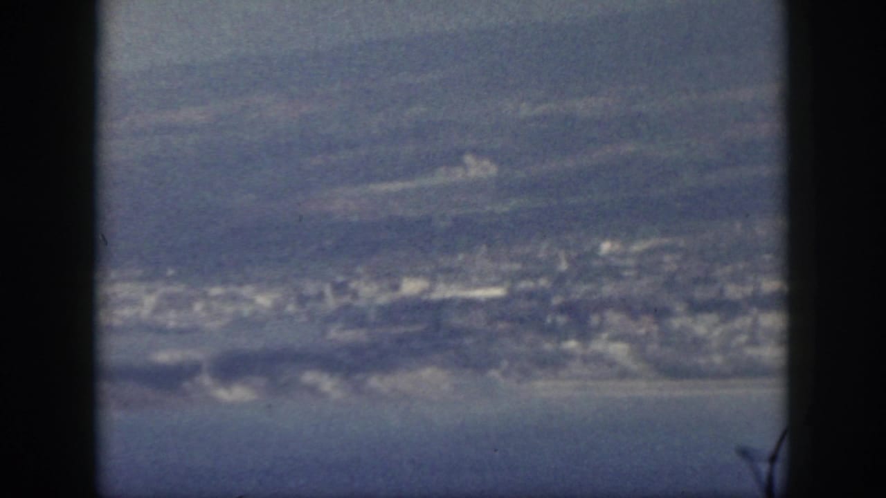 1959 Oahu home-movie still showing a shoreline settlement, blue water, and soft mountain forms in a faded 8mm frame.