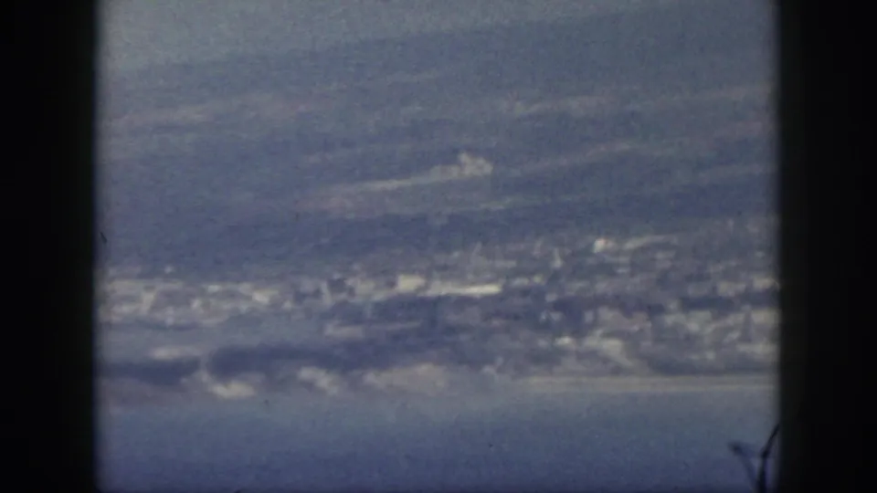 1959 Oahu home-movie still showing a shoreline settlement, blue water, and soft mountain forms in a faded 8mm frame.