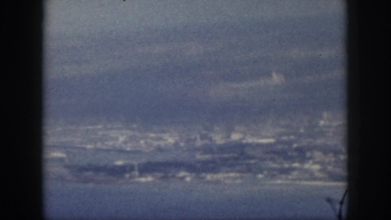 1959 Oahu frame with a distant coastal city, ocean water, and atmospheric haze seen through dark 8mm framing.