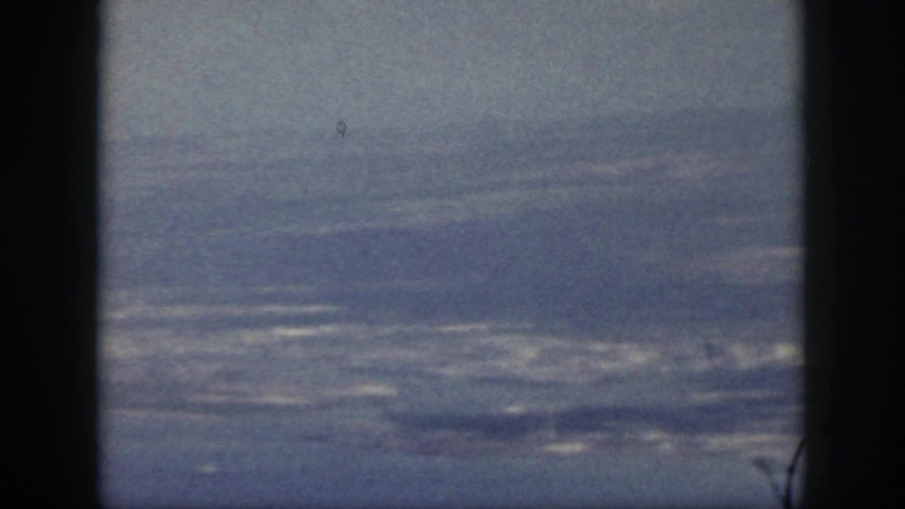 1959 Oahu home-movie frame showing a hazy coastline and ocean through a dark aircraft window divider.