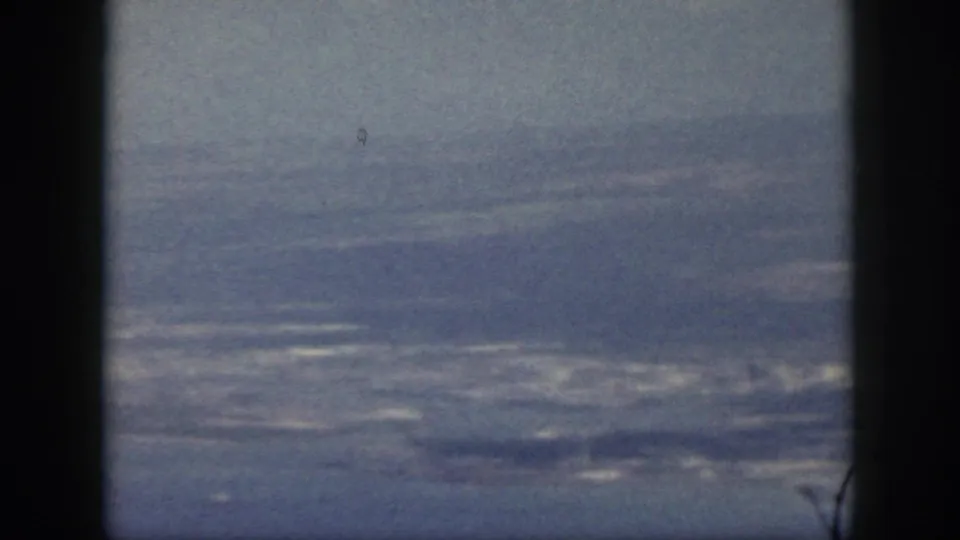 1959 Oahu home-movie frame showing a hazy coastline and ocean through a dark aircraft window divider.