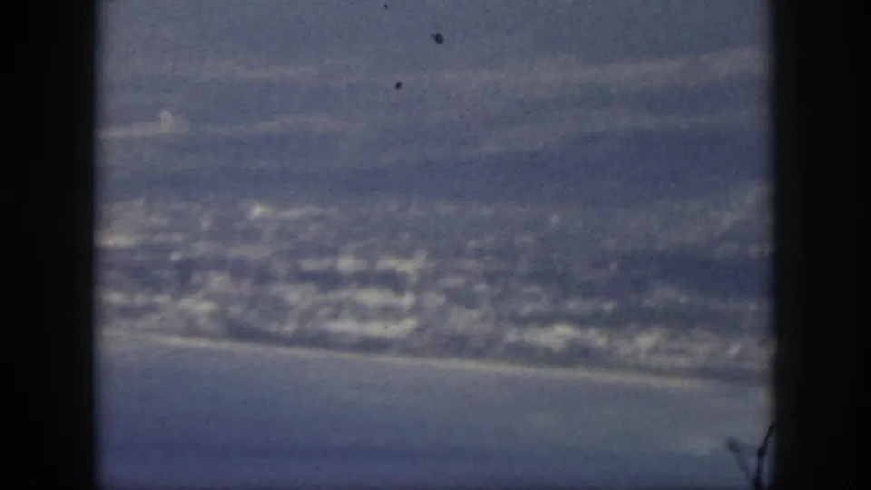1959 Oahu aerial frame with a coastal town, curved shoreline, ocean, and aircraft window edge.